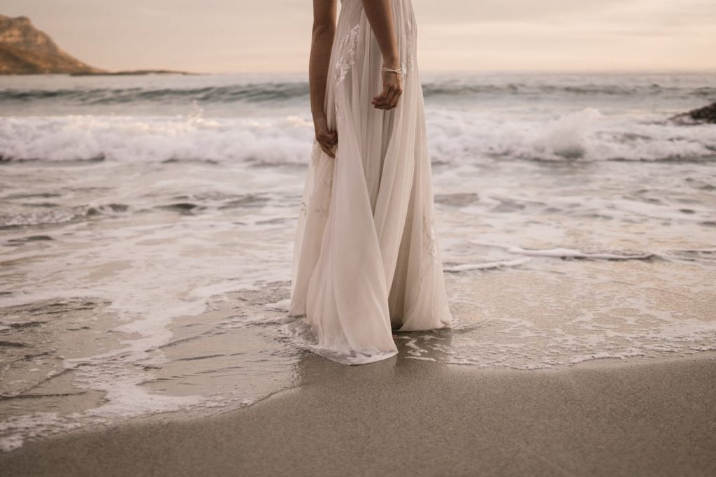 A bride in a wedding dress stands by the ocean at sunset, waves gently lapping at her feet.